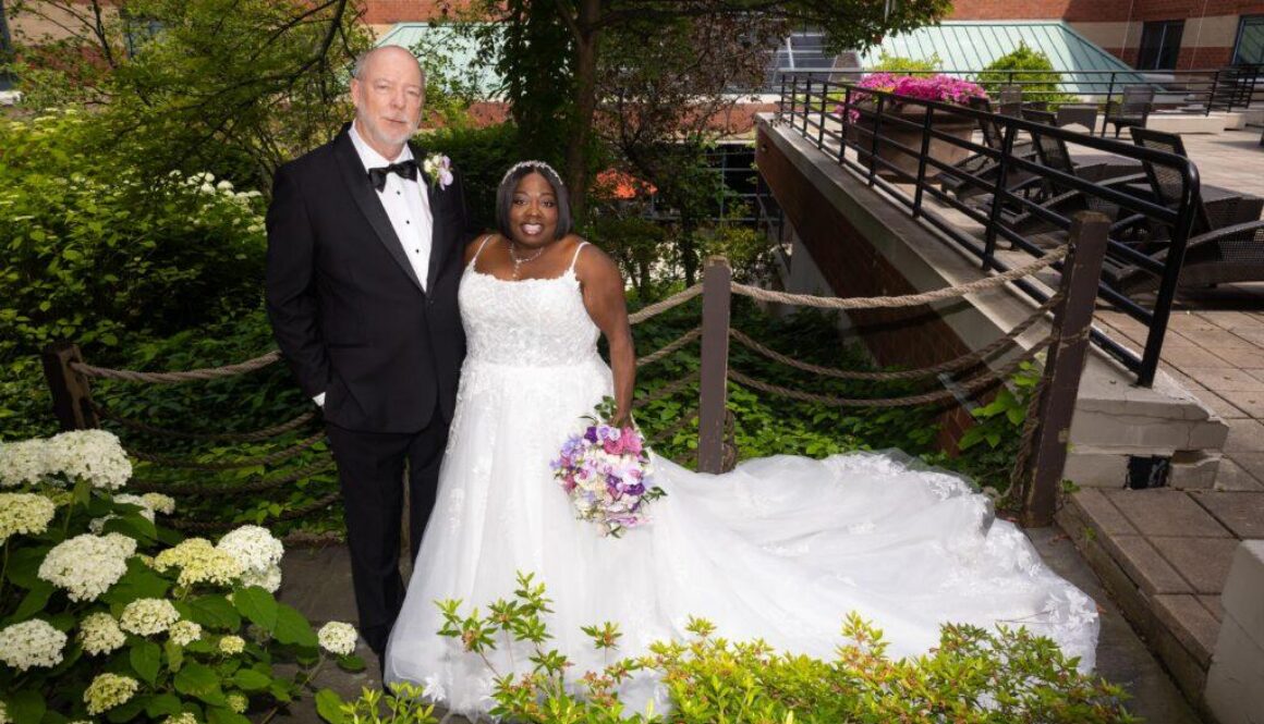 Newlyweds Catrenia and John pose outdoors in their wedding attire among lush greenery and white hydrangeas at The Baronette Renaissance.