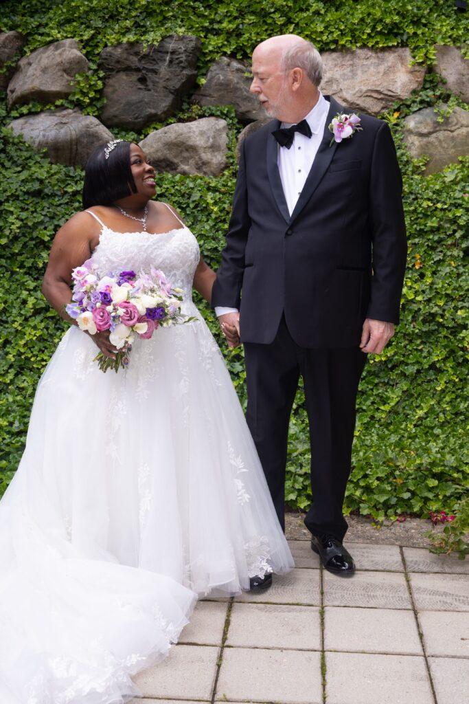 Catrenia and John hold hands and smile at each other while posing in front of a lush green ivy wall and natural stone.