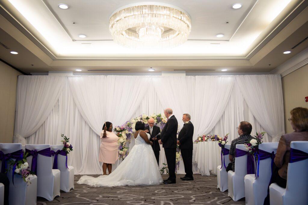 Catrenia and John stand before their officiant beneath a white and lavender floral arch during their indoor wedding ceremony.