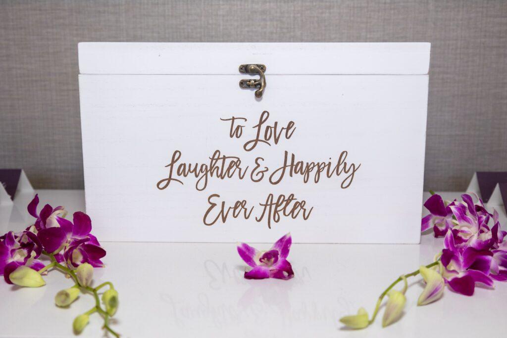 White wedding card box with gold lettering reading “To Love, Laughter & Happily Ever After,” surrounded by purple orchids.
