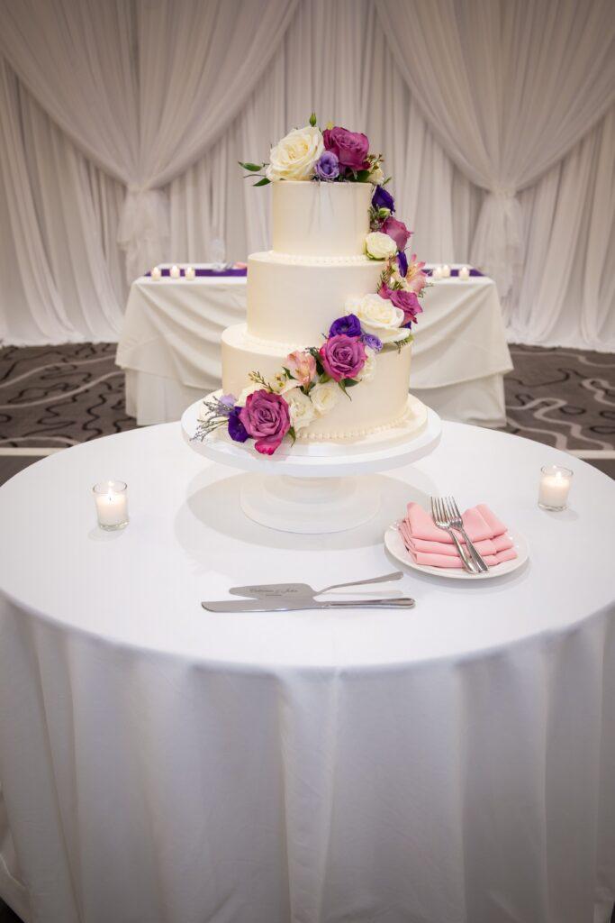 Three-tier white wedding cake decorated with cascading pink, purple, and white flowers at The Baronette Renaissance.