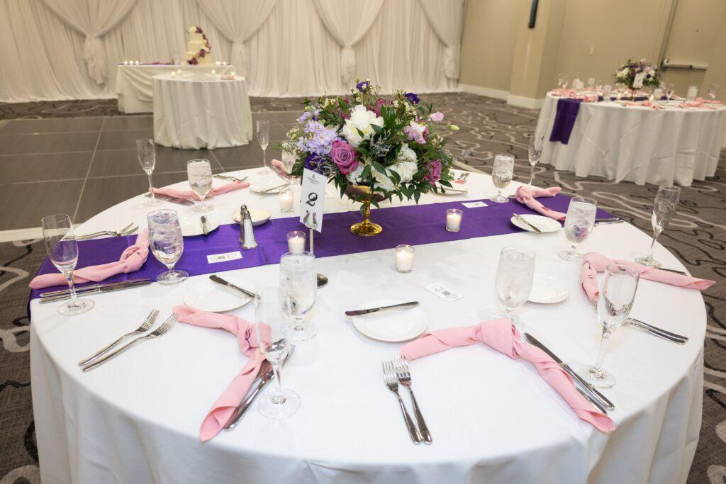 Wedding reception table set with white linens, a purple table runner, pink napkins, candles, and a colorful floral centerpiece.