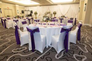 Wedding reception tables decorated with white linens, purple chair sashes, pink napkins, and purple and white floral centerpieces.