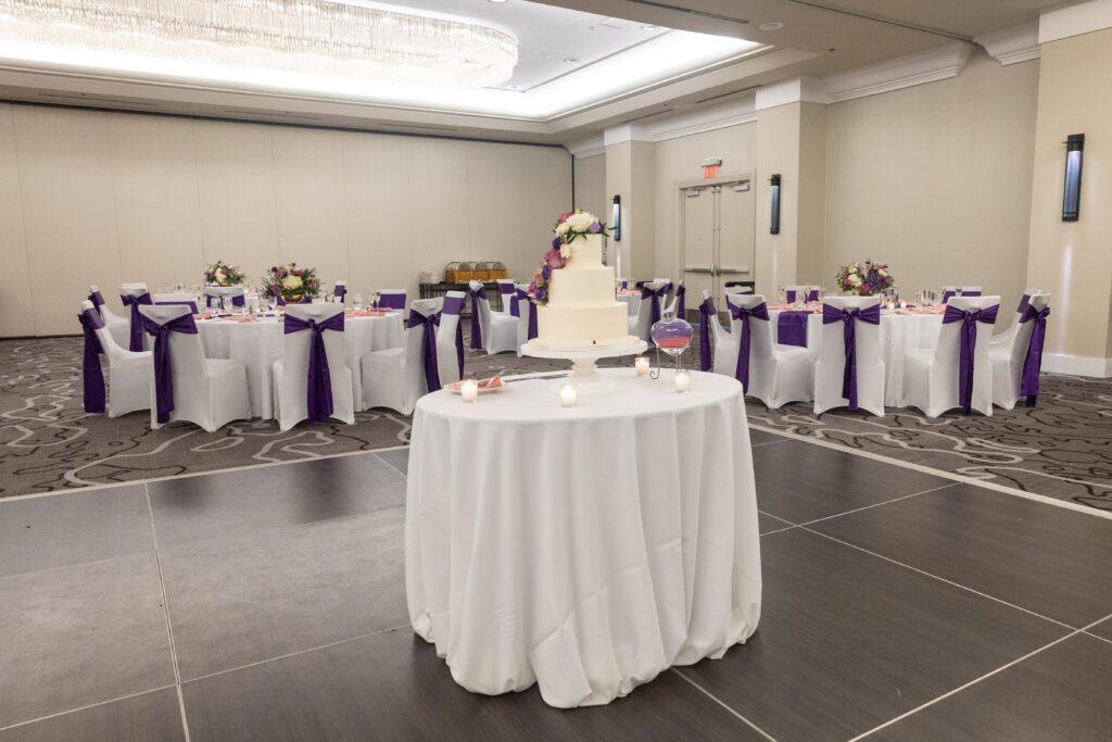 Wedding reception ballroom featuring a white floral cake on the dance floor and guest tables decorated with white linens and purple chair sashes.